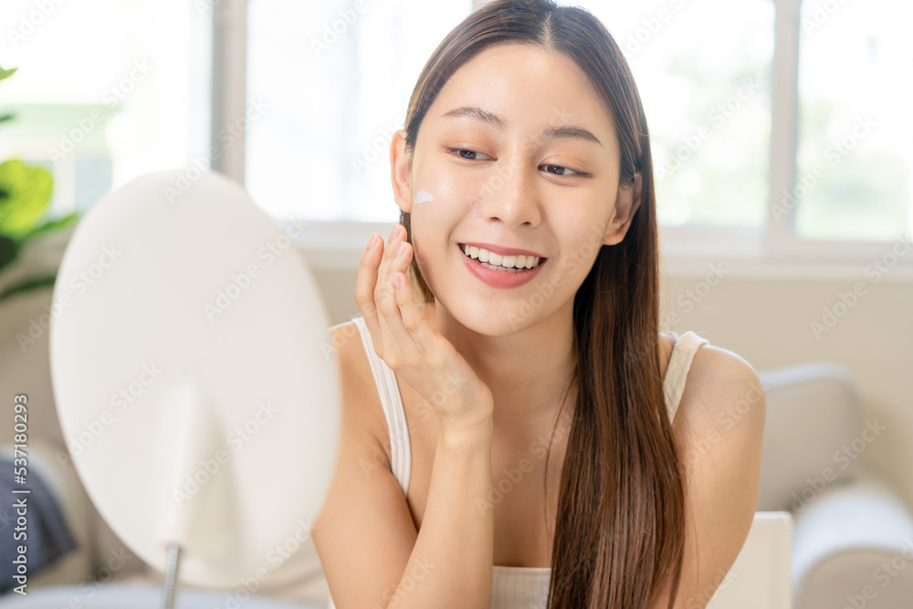 Fresh healthy skin, beautiful smile of asian young woman, girl looking at mirror, applying moisturizer on her face, putting cream treatment before makeup cosmetic routine at home. Facial Beauty. Fresh healthy skin, beautiful smile of asian young woman, girl looking at mirror, applying moisturizer on her face, putting cream treatment before makeup cosmetic routine at home. Facial Beauty.