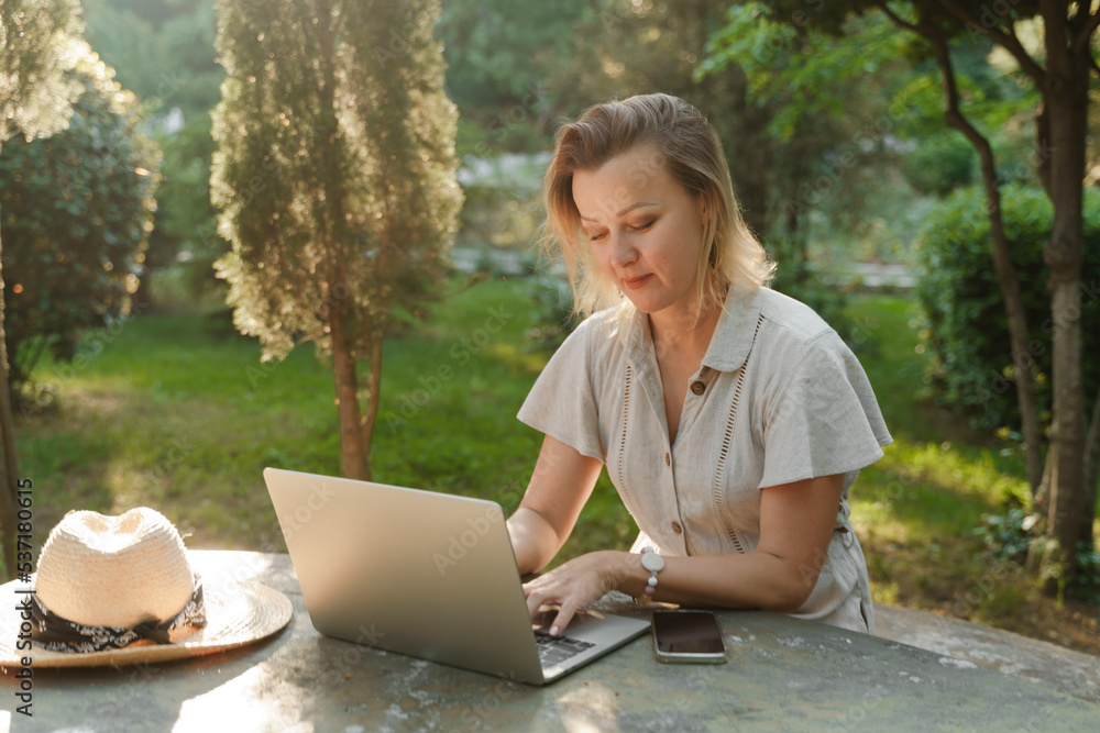 a beautiful mature woman in glasses works on a computer at a white table in nature and spends her day productively a beautiful mature woman in glasses works on a computer at a white table in nature and spends her day productively