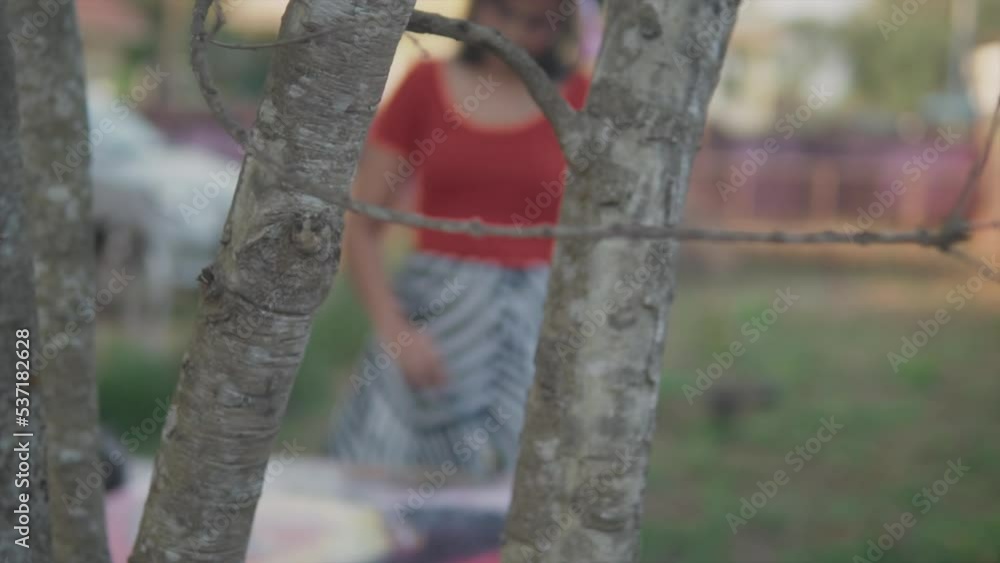 Slow panning shot through the branches of a beautiful artist dressed in red top and a black and white skirt painting a picture with acrylic paint in the garden Slow panning shot through the branches of a beautiful artist dressed in red top and a black and white skirt painting a picture with acrylic paint in the garden