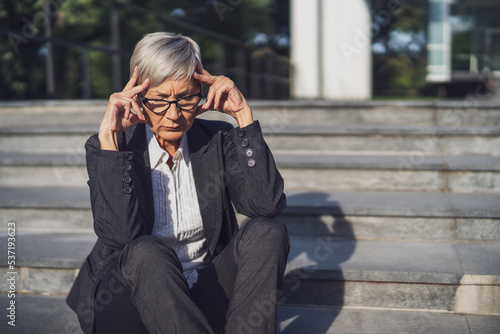 Senior businesswoman sitting in front of company building. She is tired and having headache.