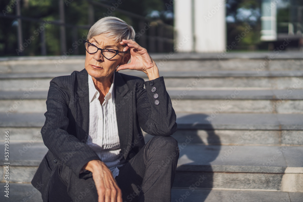 Senior businesswoman sitting in front of company building. She is thoughtful and worried. Senior businesswoman sitting in front of company building. She is thoughtful and worried.