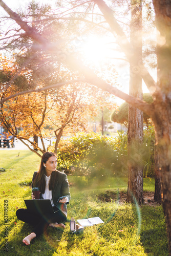 Barefoot young caucasian woman wearing jacket sitting on grass with laptop and single-use cup of coffee, searching information for her job. Young creative