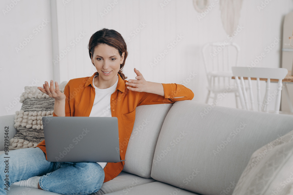 Naklejka premium Surprised girl chatting by video call at laptop, astonished with good news, sitting on couch at home