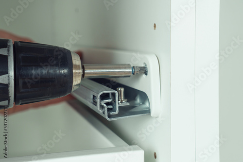 Фотография Male hands hold cordless electric screwdriver, screw to fix rail, assembling drawer of white dresser or wardrobe