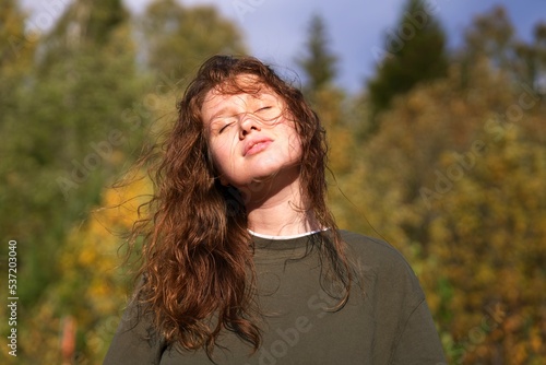 Portrait of happy relaxed young beautiful woman, calm girl is relaxing, meditating breathing deep deeply fresh autumn air at sunny day in forest with eyes closed, smile. Sunshine, natural background