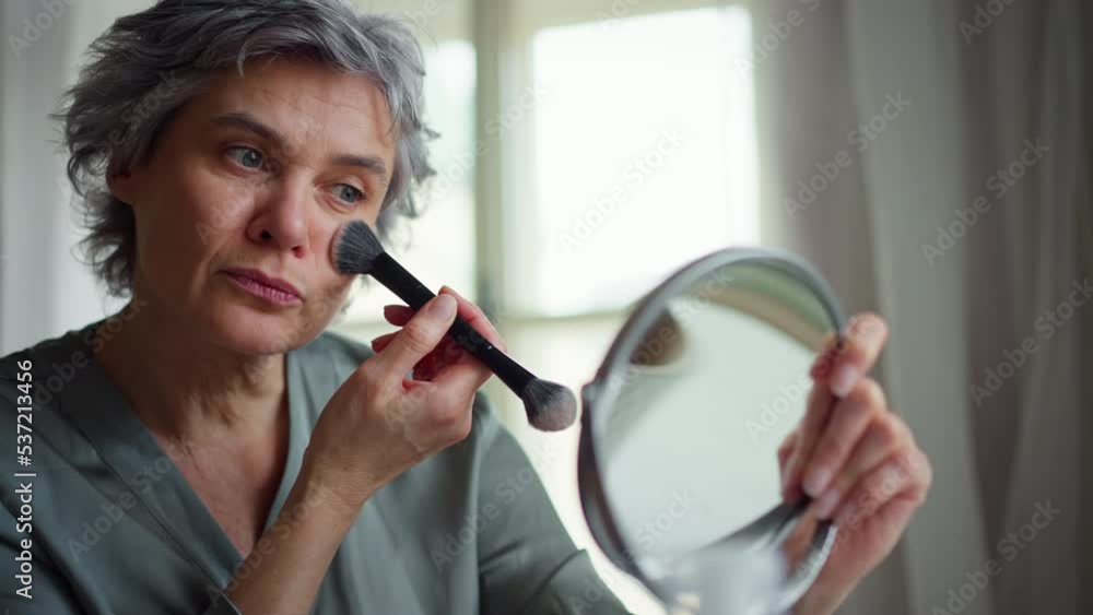 Attractive mature woman at home, sitting at cosmetic table, putting in powder with brush, observing herself in mirror. Attractive mature woman at home, sitting at cosmetic table, putting in powder with brush, observing herself in mirror.