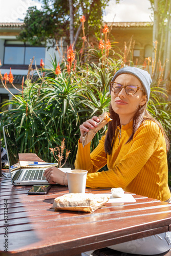 Hispanic female student sitting on college campus in front of her computer and notebook taking a break while eating a snack
