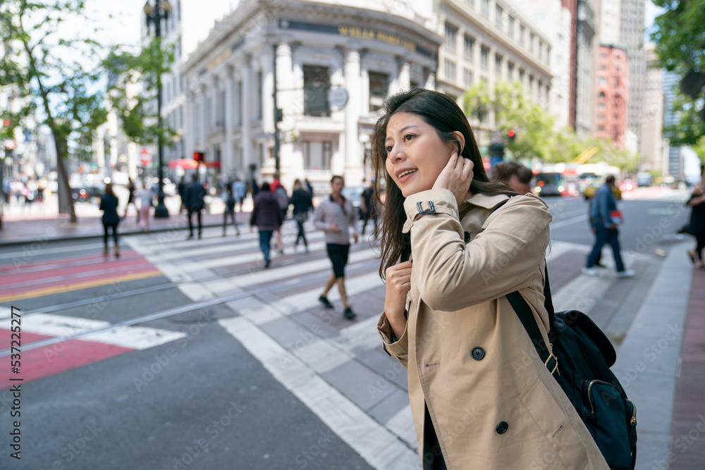 asian Korean female businessperson tucking hair with smile and looking into distance while waiting for bus near crosswalk in san Francisco California usa. people are crossing road at background