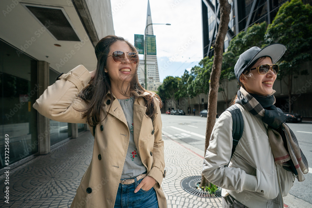 Naklejka premium happy asian businesswoman holding her messy hair while walking with her colleague on street during lunch break on windy day in san Francisco with Transamerica Pyramid at background