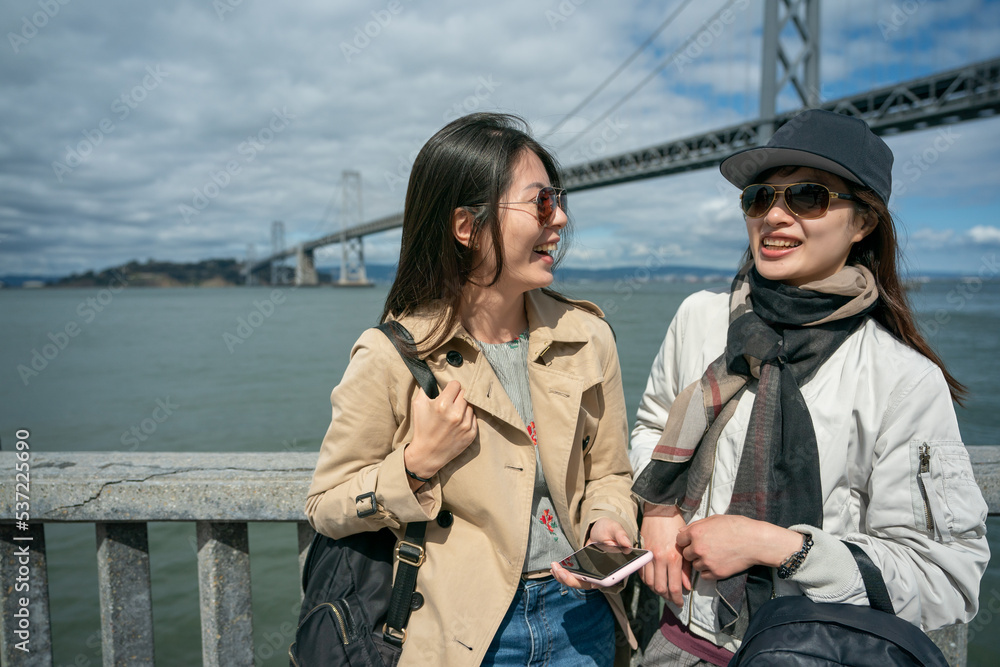 Naklejka premium two happy asian Korean girl friends wearing sunglasses talking happily near railing with Oakland bay bridge at background while traveling to San Francisco together in spring