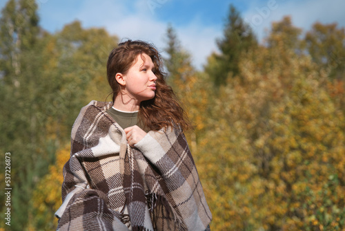 portrait of beautiful cozy girl, young woman standing in golden autumn park or forest covering herself in blanket, plaid looking to the side. Copy space, place for text.