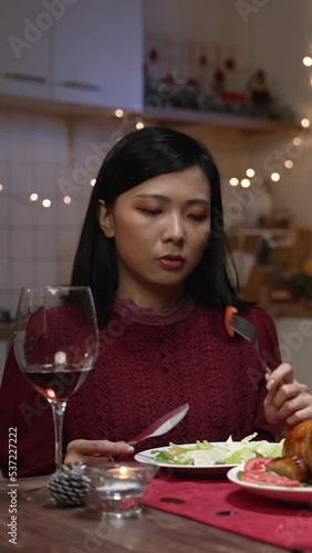 Vertical Screen: annoyed asian wife feeling upset while eating Christmas dinner because her husband is playing with cell phone and ignoring her at table in decorated dining room