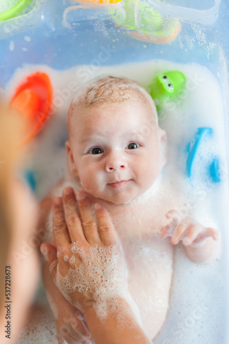 Baby bath time. Close-up detail view of mother bathing cute little peaceful baby in tub with water and bubbles lather.