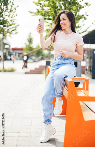 Beautiful cheerful smiling woman with earphones listening music on her mobile phone sitting outdoors at the street while enjoying.