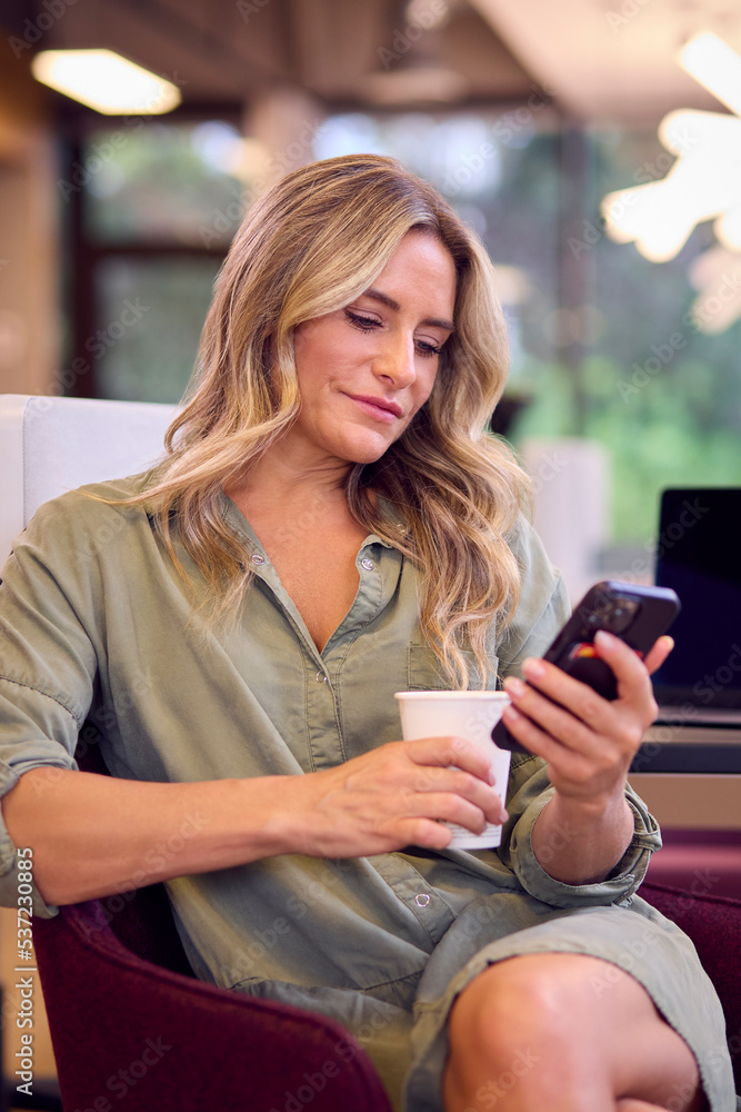 Mature Businesswoman Sitting At Desk In Office With Takeaway Drink Looking At Mobile Phone Mature Businesswoman Sitting At Desk In Office With Takeaway Drink Looking At Mobile Phone