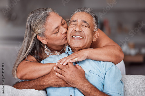 Happy senior couple hug in home, retirement lifestyle together in Mexico and relax on sofa. Romantic marriage life, love bonding time and elderly woman kissing latino husband on cheek in living room