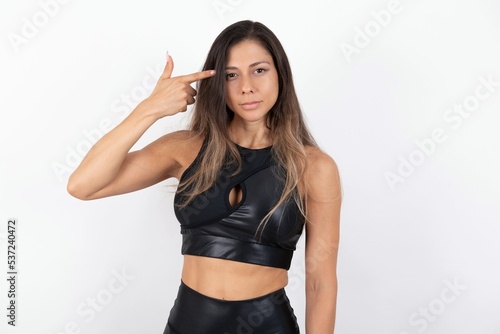Fototapeta Naklejka Na Ścianę i Meble -  Unhappy young beautiful woman wearing sportswear over white background makes suicide gesture and imitates gun with hand, curves lips keeps two fingers on temple, shoots, being tired of everything,