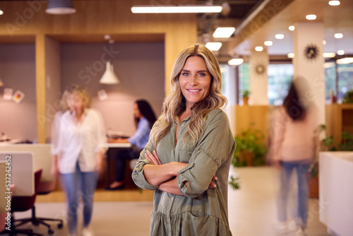 Portrait Of Businesswoman Standing In Busy Open Plan Office With Motion Blur Colleagues Behind
