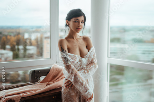 Sexy girl in a white translucent tunic sits sewing near the window.Beautiful female body shines through the fabric