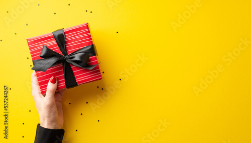 Black friday concept. First person top view photo of female hand in black shirt holding red giftbox with ribbon bow over confetti on isolated yellow background with copyspace