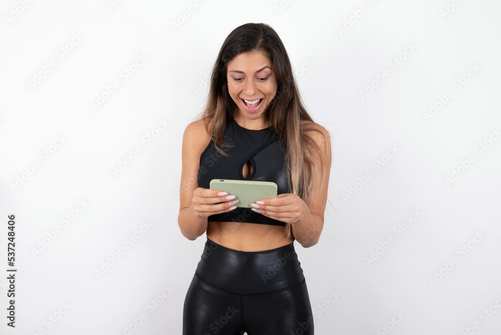 Portrait of an excited young beautiful woman wearing sportswear over white background playing games on mobile phone. Portrait of an excited young beautiful woman wearing sportswear over white background playing games on mobile phone.