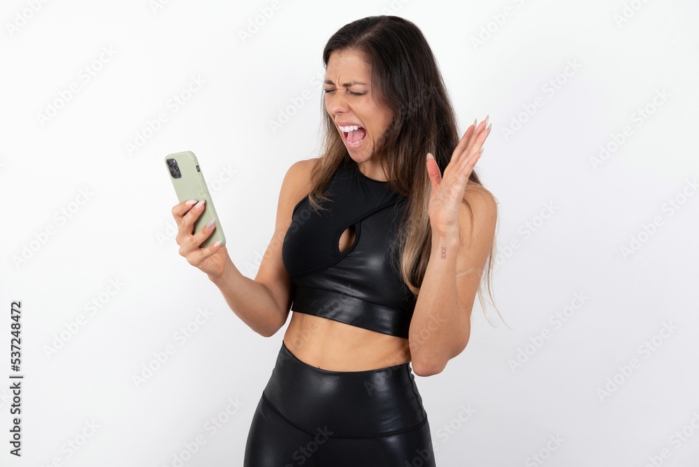 Angry young beautiful woman wearing sportswear over white background screaming on the phone, having an argument with an employee. Troubles at work. Angry young beautiful woman wearing sportswear over white background screaming on the phone, having an argument with an employee. Troubles at work.