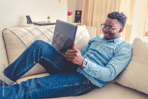 Young Afro-american man sitting on couch with laptop, working remotely, using apps.