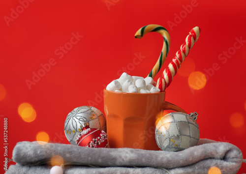 Xmas composition. Cozy mug with cacao, marshmallows, licorice candies and Christmas baubles on stack of warm sweaters on festive red background. Winter textured layout with copy space