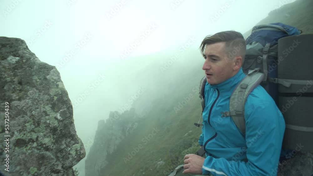 A boy tourist takes a selfie in the mountains. Mystical landscape of peaks in the haze and a happy man. Active recreation and hiking in the mountains with a backpack. High quality 4k footage A boy tourist takes a selfie in the mountains. Mystical landscape of peaks in the haze and a happy man. Active recreation and hiking in the mountains with a backpack. High quality 4k footage