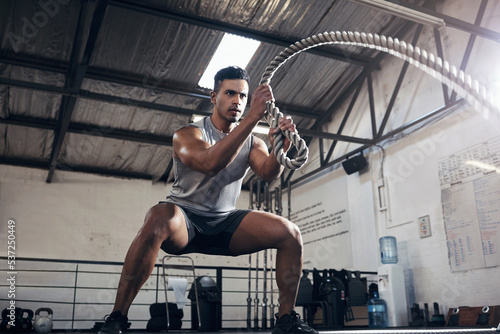 Health, gym and battle rope workout with man at a fitness center, cardio and muscle training. Power, energy and exercise by muscular athlete focused on intense movement, strength and endurance photo