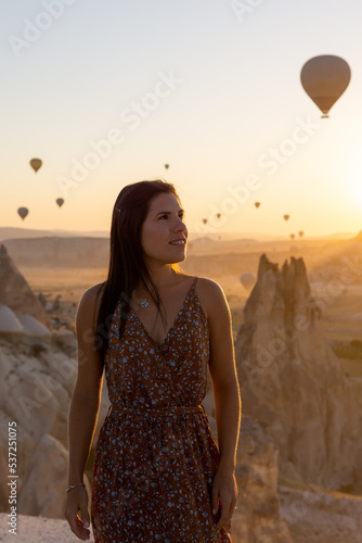 Portrait of a happy young woman with the silhouette of hot air balloons flying at sunrise in the background.