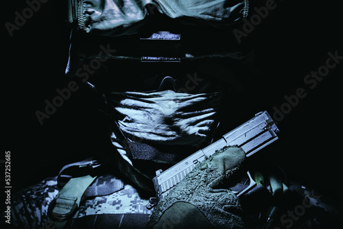 Special operations forces soldier in combat helmet with hidden behind balaclava and dark glasses face posing with sidearm service pistol in hand. Tone, high contrast, cropped on black background