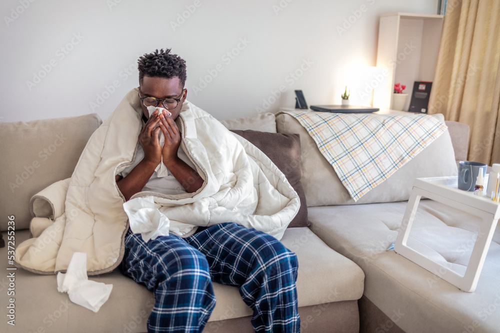 Cropped shot of a young man suffering with flu while sitting wrapped in a blanket on the sofa at home. Cropped shot of a young man suffering with flu while sitting wrapped in a blanket on the sofa at home.