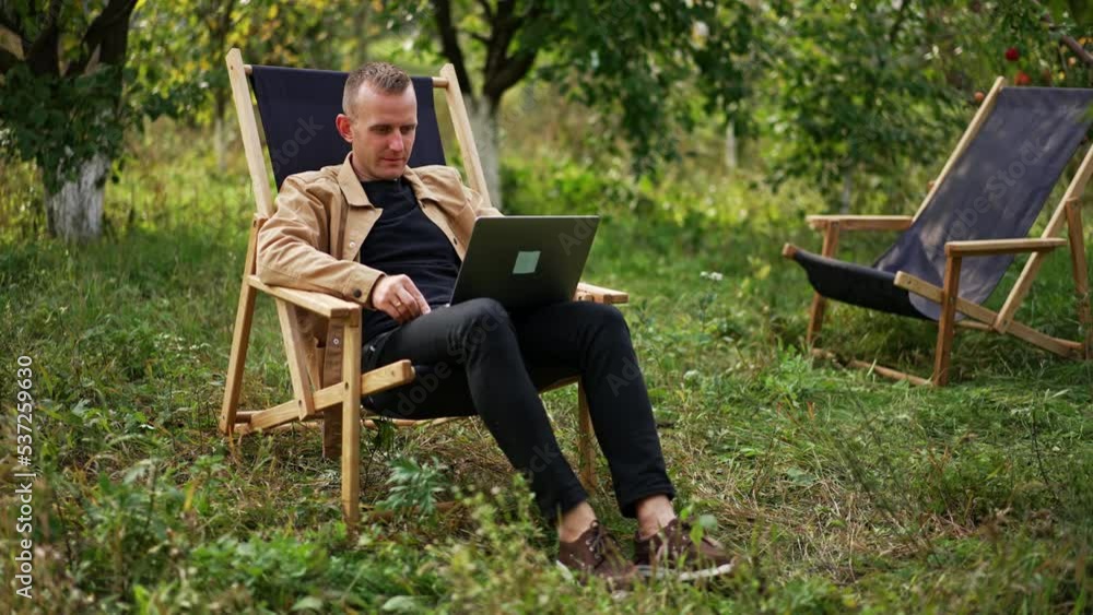 Calm relaxed freelancing man sitting in apple garden with laptop on his knees. Caucasian male working remotely online from nature. Calm relaxed freelancing man sitting in apple garden with laptop on his knees. Caucasian male working remotely online from nature.