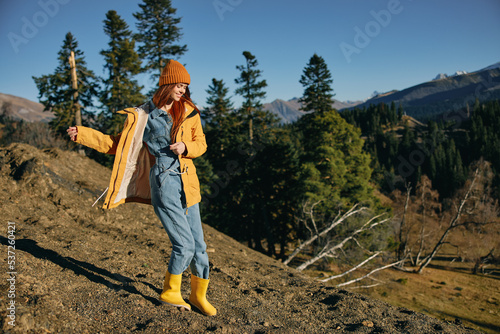 Woman traveling to the mountains in the fall on a nature hike smile and happiness in a yellow cape with red hair full-length stands against the backdrop of trees and mountains