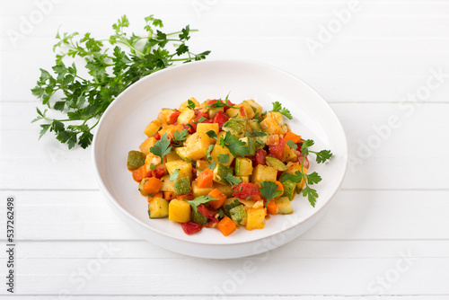 Vegetable stew with zucchini, carrots, potatoes and parsley on a white plate on a light gray background