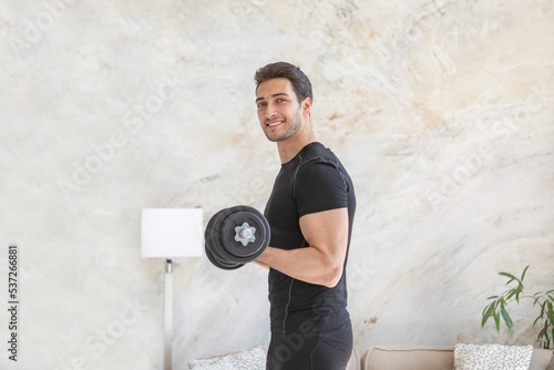 Foto Side view of young handsome man in black sportswear doing biceps curl with dumbbells in living room at home