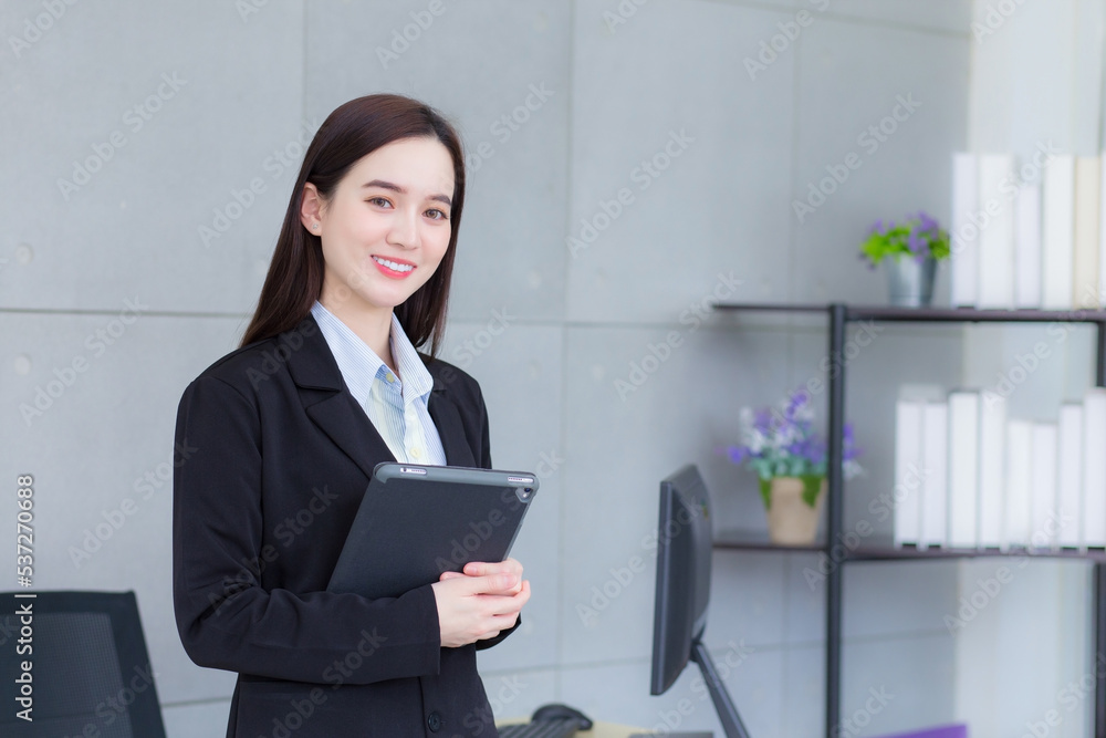 Asian professional business young woman in black suit smiles happily stand with confidence and look at the camera while she works and holds tablet in office. Asian professional business young woman in black suit smiles happily stand with confidence and look at the camera while she works and holds tablet in office.