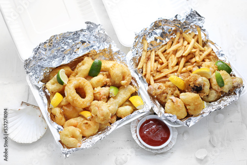 Deep fried calamari, squid rings and shrimps in tempura, with fries, in take-away carton boxes, on a white background. 