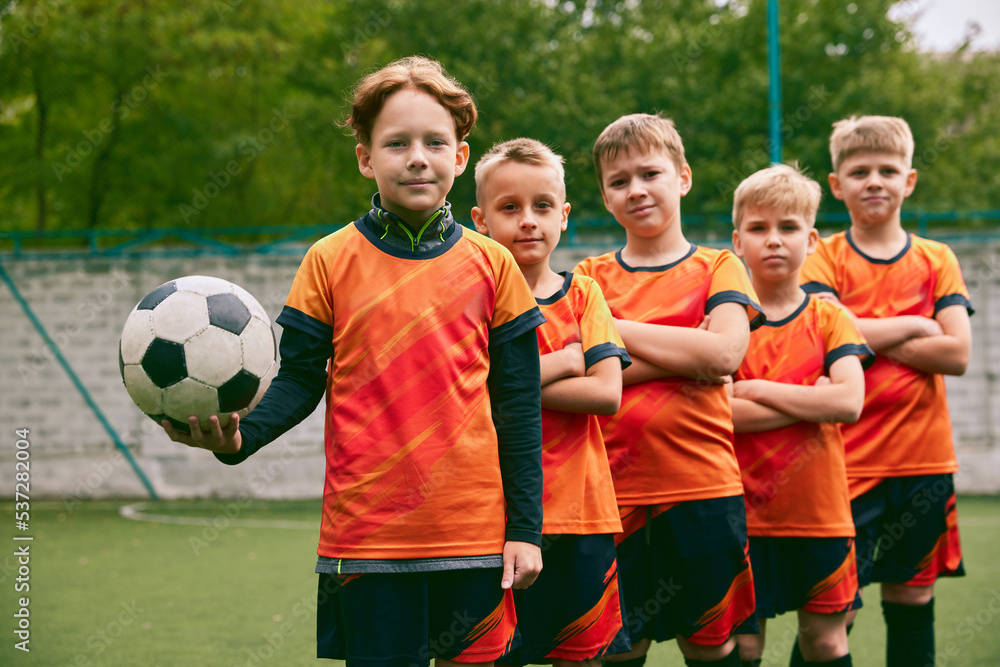 Fototapeta premium Teammates. Athletic boys in junior soccer team standing together at grass sport field. Football players in orange-black kits and boots.