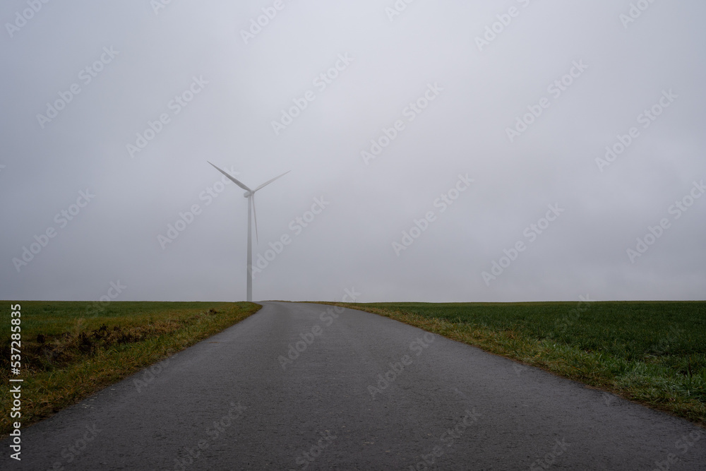 Asphalt walkway in the countryside with a white wind turbine on a foggy morning Asphalt walkway in the countryside with a white wind turbine on a foggy morning