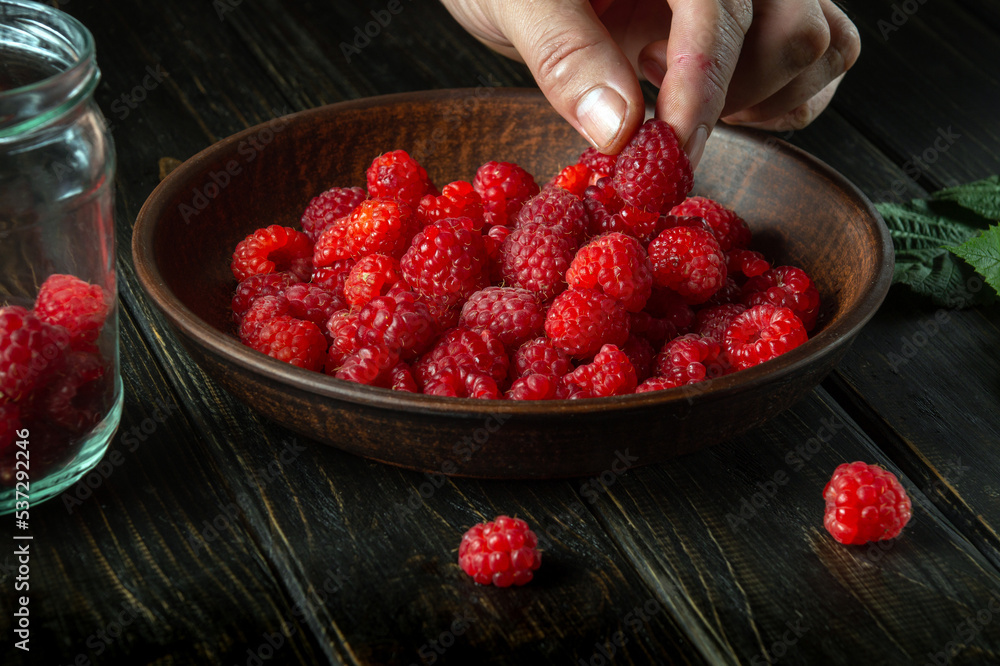 The cook sorts through fresh ripe raspberries before preparing a sweet compote in a jar. Close-up of chef hands while working The cook sorts through fresh ripe raspberries before preparing a sweet compote in a jar. Close-up of chef hands while working