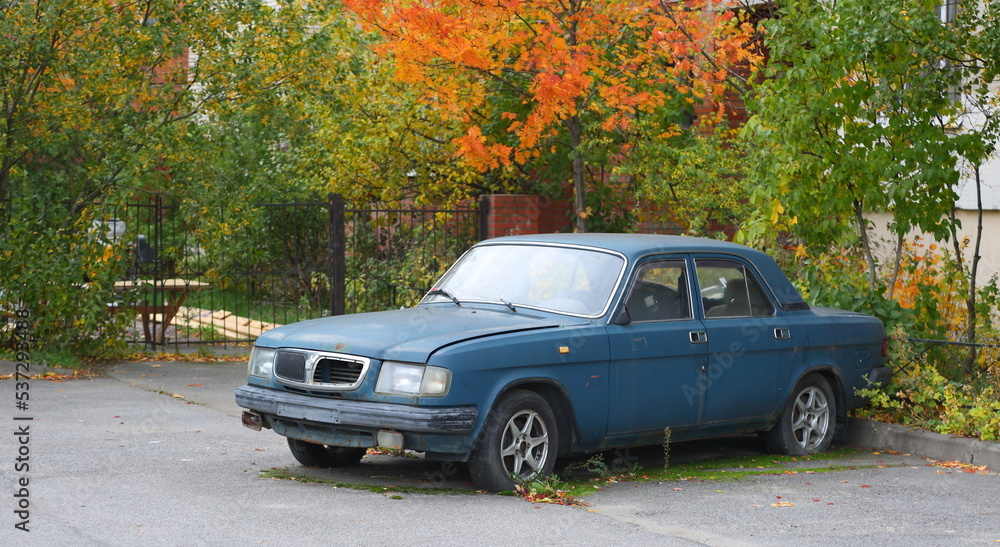 An old rusty blue car is parked near the autumn bush, Kollontai Street, St. Petersburg, Russia, October 2022