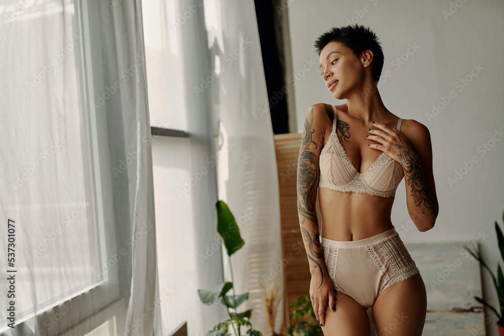 Young tattooed woman in lace lingerie looking at window at home. - obrazy, fototapety, plakaty 