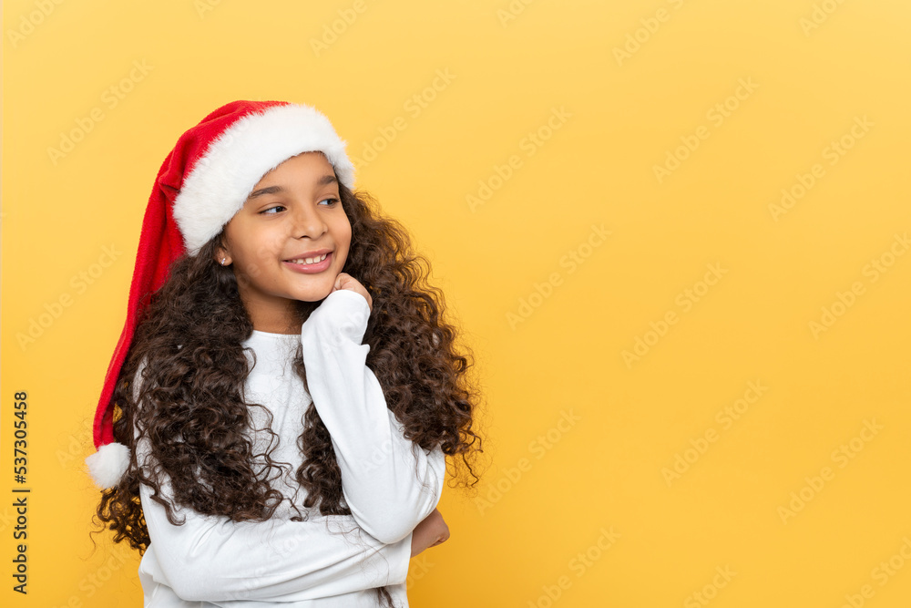 Dark skinned young girl with long curly hair in a Santa Claus hat on a yellow background Dark skinned young girl with long curly hair in a Santa Claus hat on a yellow background