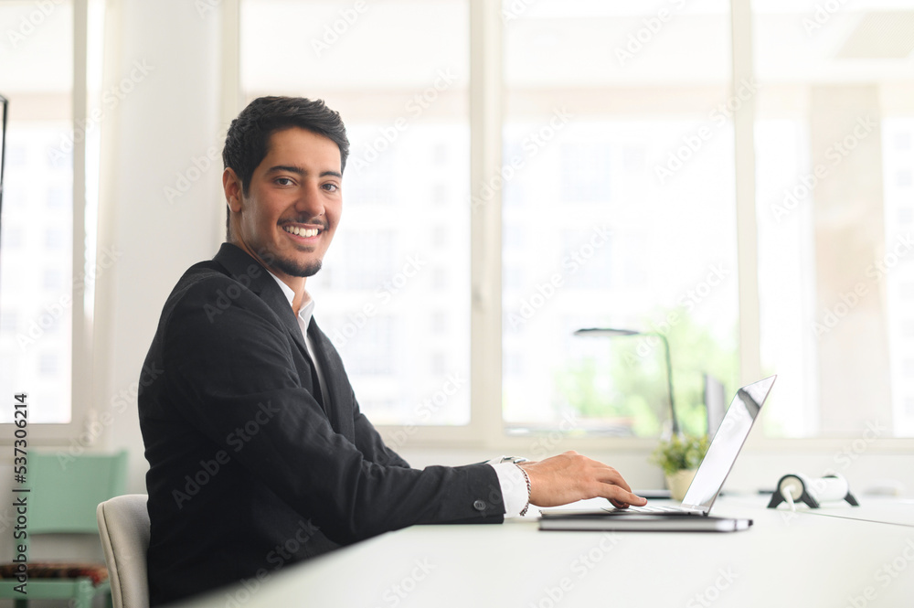Cheerful latin male freelancer or office worker is using laptop computer sitting at the desk. Friendly and optimistic eastern dark-haired guy in formal suit looks at camera and smiles, side view Cheerful latin male freelancer or office worker is using laptop computer sitting at the desk. Friendly and optimistic eastern dark-haired guy in formal suit looks at camera and smiles, side view