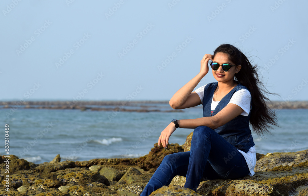 Young lady wearing jeans and t-shirt sitting on the coral of a sea beach. Beautiful stylish woman enjoying the beauty of a nature.