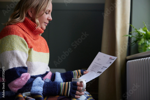 Woman In Gloves With Hot Drink And Bill Trying To Keep Warm By Radiator During Cost Of Living Energy Crisis photo