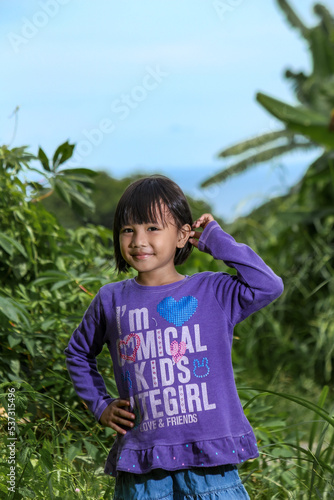 Happy little girl posing outdoors with a smile, wearing a purple shirt and denim skirt, standing in nature with green plants and blue sky background.