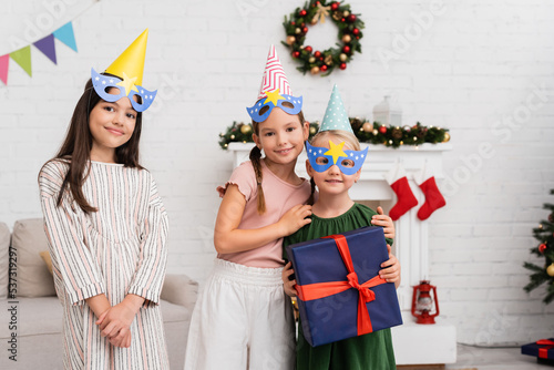 Smiling girls in party caps and masks hugging friend with present near christmas decor at home.